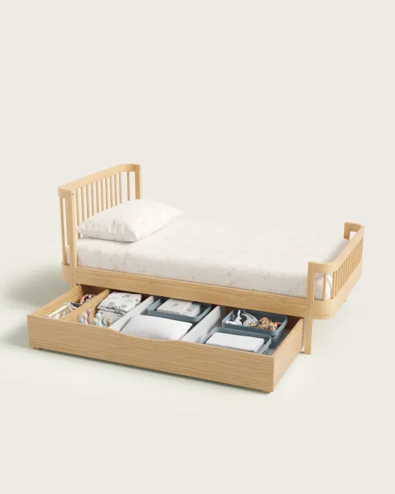 A wooden single bed with white bedding and an under-bed storage drawer containing toys, books, and baskets, against a plain background.