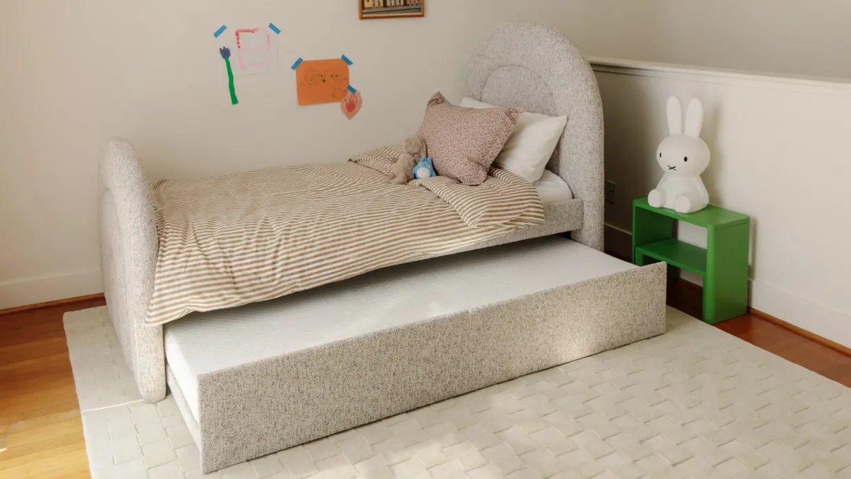 A neatly made bed with striped bedding and a pull-out trundle, children’s drawings on the wall, and a green nightstand with a white bunny lamp.