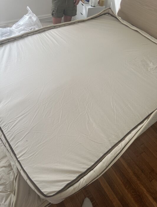 A person stands near an unmade bed with a large, cream-colored mattress cover, partially unzipped, on a wooden floor in a bright room.