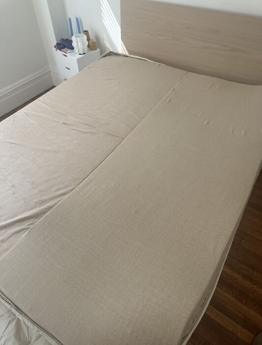 A beige bed with a visible crease down the center, next to a wooden headboard and a small white nightstand on a wooden floor.