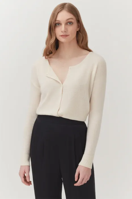 Woman stands against a plain background, wearing a cream-colored long-sleeve sweater with a few top buttons undone and high-waisted black pants.
