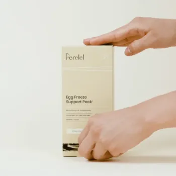 A person opens a beige box labeled “Perelel Egg Freeze Support Pack” multivitamin and supplement. The background is plain and light-colored.