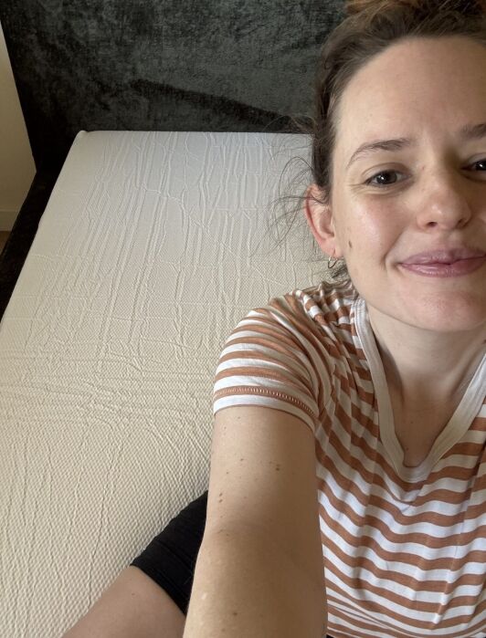 Person wearing a striped shirt sits on the edge of a bare mattress, smiling at the camera. The background includes a dark headboard.