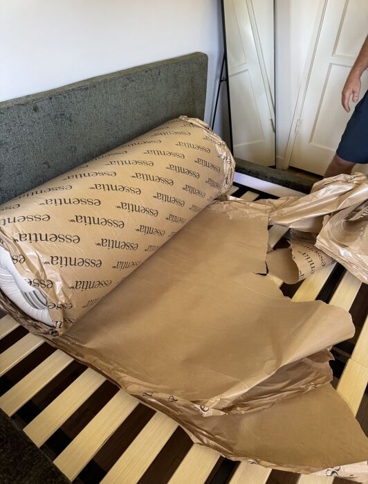 A rolled-up mattress in branded packaging is partially unwrapped and resting on a slatted bed frame, with torn brown paper nearby.