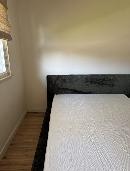 A neatly made bed with a white sheet is placed against a plain wall next to a window with blinds in a minimally furnished room with wooden flooring.