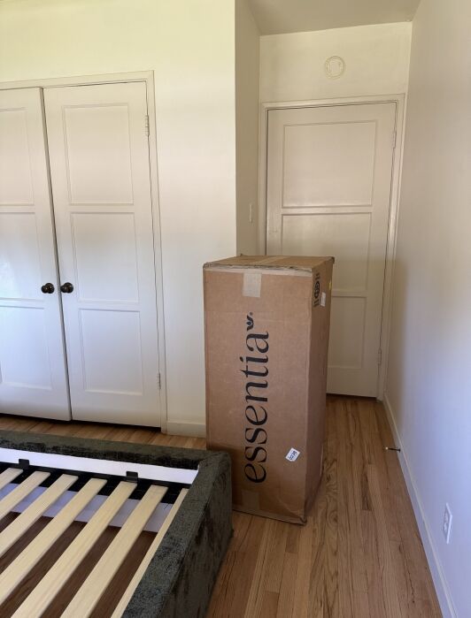 A cardboard box labeled “essentia” stands upright in a bedroom with a wooden floor, next to an unassembled bed frame and closed doors.