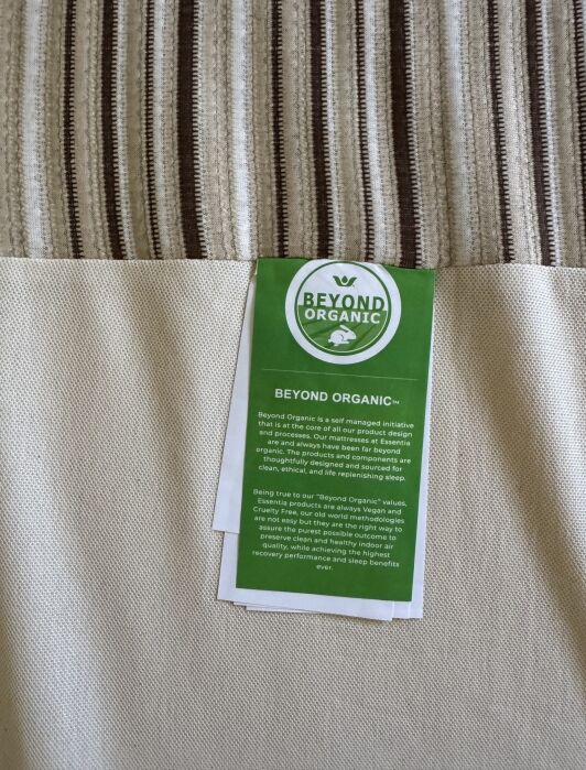 A green “Beyond Organic” label with white text is attached to beige and striped fabric, describing the initiative’s commitment to organic, ethical, and sustainable practices.