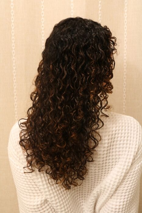 A person with long, dark, tightly curled hair is shown from the back, wearing a textured white top against a beige patterned background.