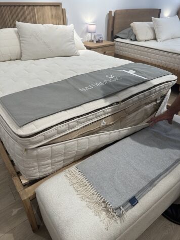 A hand lifts a gray cover to reveal the unzipped side of a mattress in a showroom, next to a bench with a folded fringed throw. Other beds and lamps are visible in the background.
