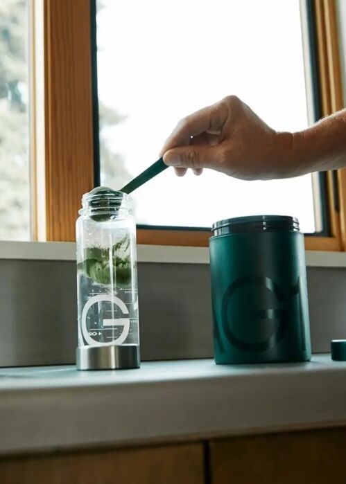 A hand adds powdered supplement from a green container into a clear water bottle on a kitchen counter near a window.