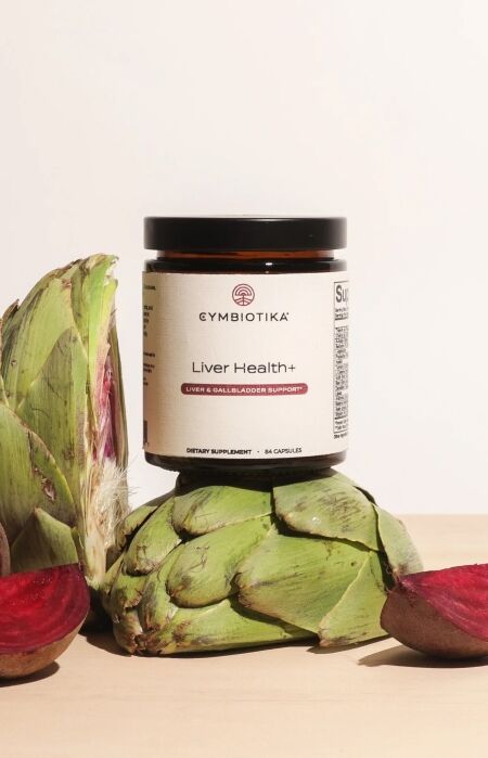 A jar of Cymbiotika Liver Health+ supplement is displayed between artichokes and sliced beets on a beige surface.