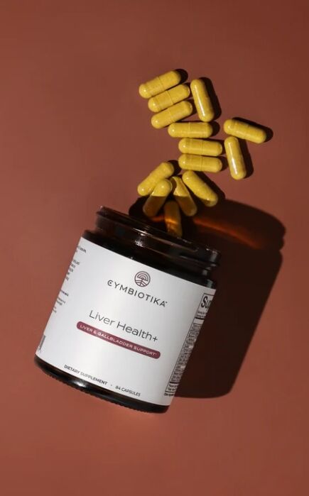 A brown bottle labeled “Liver Health+” lies on its side with yellow capsules spilling out onto a brown surface.