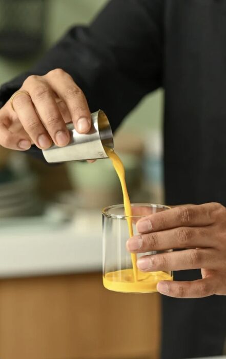 A person pours a thick yellow liquid from a small metal cup into a clear glass while holding both containers with their hands.