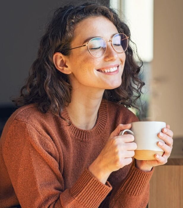 A woman with curly brown hair and glasses, wearing a rust sweater, sits indoors smiling with eyes closed while holding a white mug.