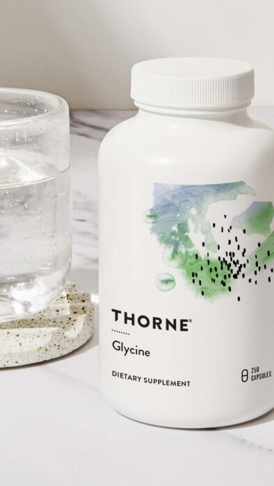 A white bottle labeled “Thorne Glycine Dietary Supplement” sits next to a glass of water on a marble surface.