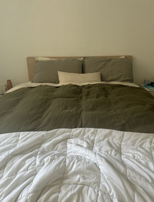 A neatly made bed with olive green bedding, two green pillows, a beige pillow, and a white quilted blanket at the foot, against a light-colored wall.
