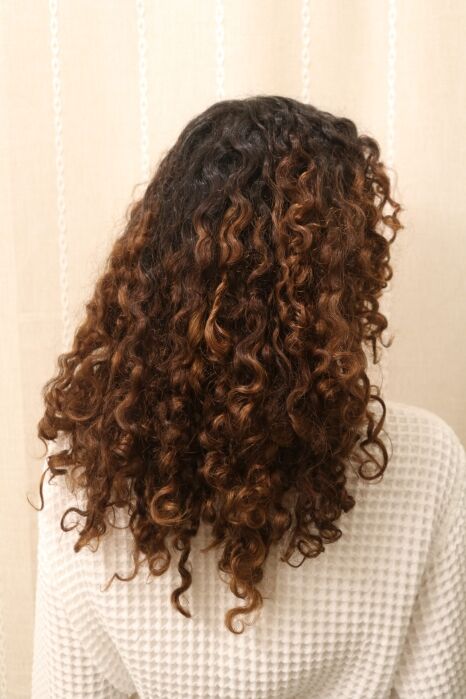 Person with shoulder-length, dark brown curly hair featuring lighter brown highlights, wearing a textured white top, shown from behind against a beige background.
