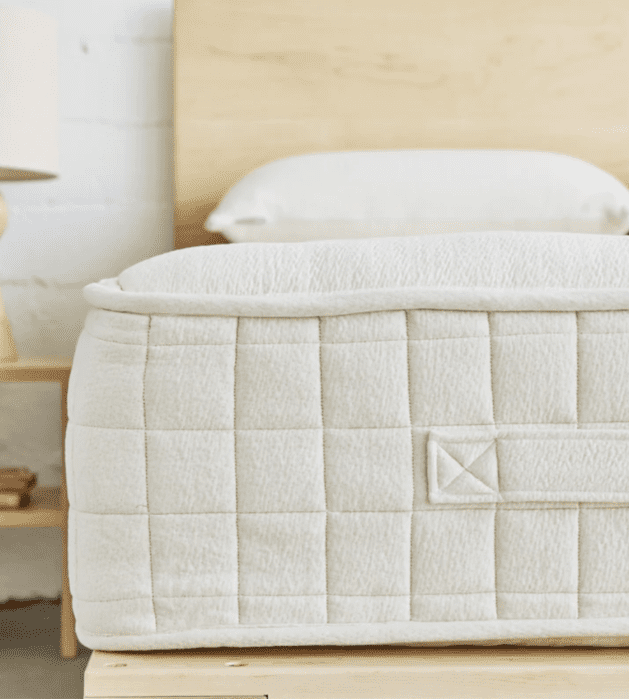 Close-up of a quilted mattress with a fabric handle, placed on a wooden bed frame. A pillow and headboard are visible in the background.
