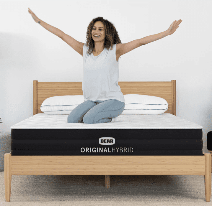 Woman with curly hair sits smiling on a Bear Original Hybrid mattress with arms raised, on a wooden bed frame in a minimal bedroom.