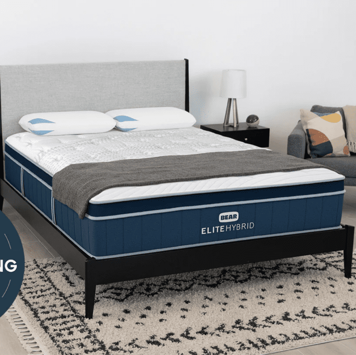 A neatly made bed with a blue Bear Elite Hybrid mattress, two pillows, a gray blanket, and a modern bedside table in a tidy, contemporary bedroom.