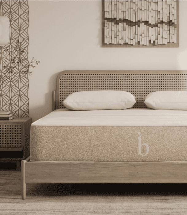 A modern bedroom features a wooden bed with a tan mattress, two cream pillows, a nightstand, geometric wallpaper, and a wall art piece above the headboard.