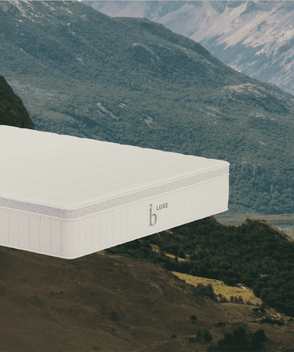 A white mattress with “LUXE” and a lowercase “b” logo floats in front of a scenic mountain landscape with green valleys.