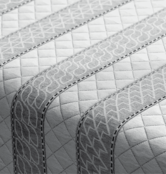 A close-up of a quilted fabric with alternating diagonal lines and geometric patterns in shades of grey.