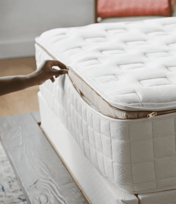 A hand unzips the top layer of a white, quilted mattress to reveal an inner layer, showing the mattress’s removable or adjustable feature.
