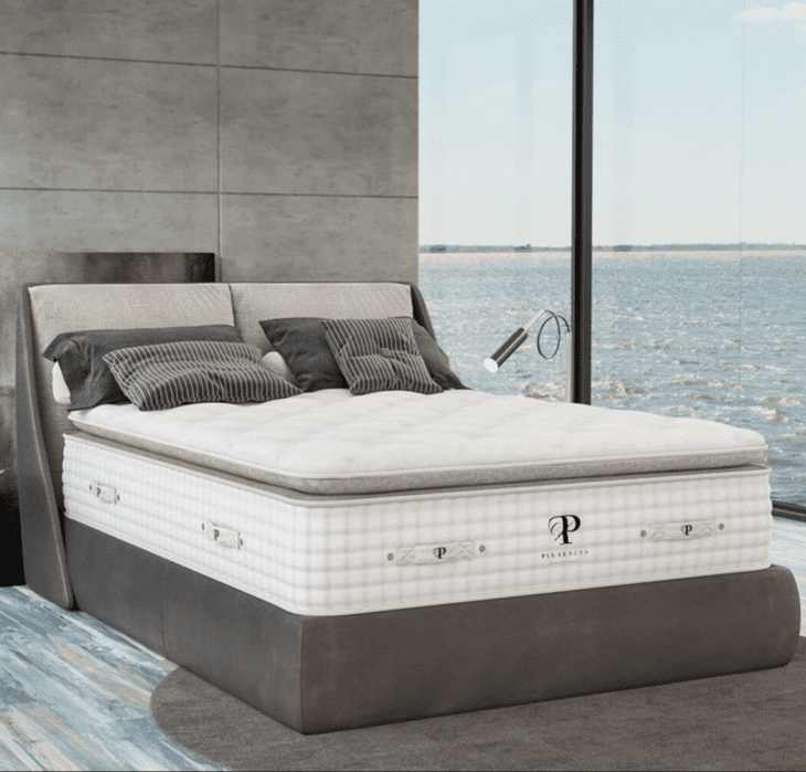 Modern bed with pillows and a plush mattress in a room with large glass windows overlooking a body of water.