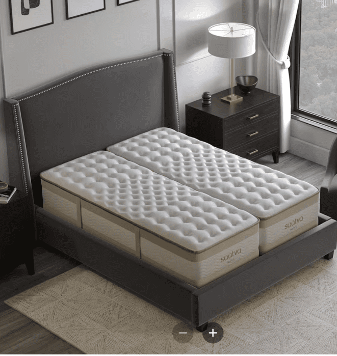 A modern bedroom with a gray upholstered bed frame holding two twin Saatva mattresses side by side, next to a nightstand, lamp, and window.