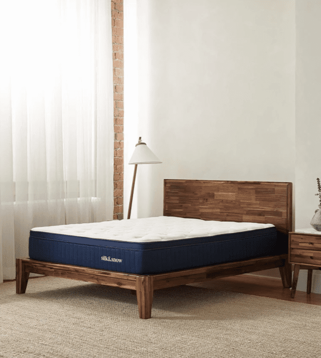 A wooden bed frame with a mattress sits in a minimalist bedroom with a nightstand, floor lamp, white curtains, and woven rug on the floor.