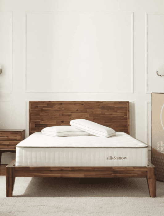 A wooden bed frame with a mattress and two pillows sits in a minimal bedroom with white walls and a nightstand. A large box labeled “silk & snow” stands next to the bed.