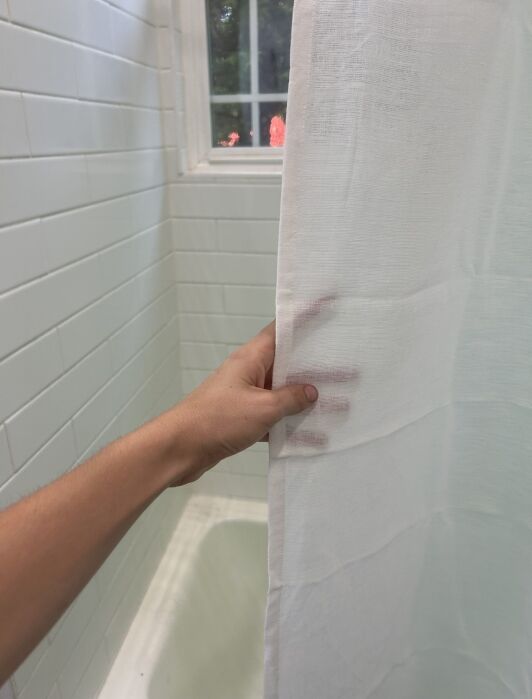A hand holds a white shower curtain partially open in a tiled bathroom with a window above the bathtub.