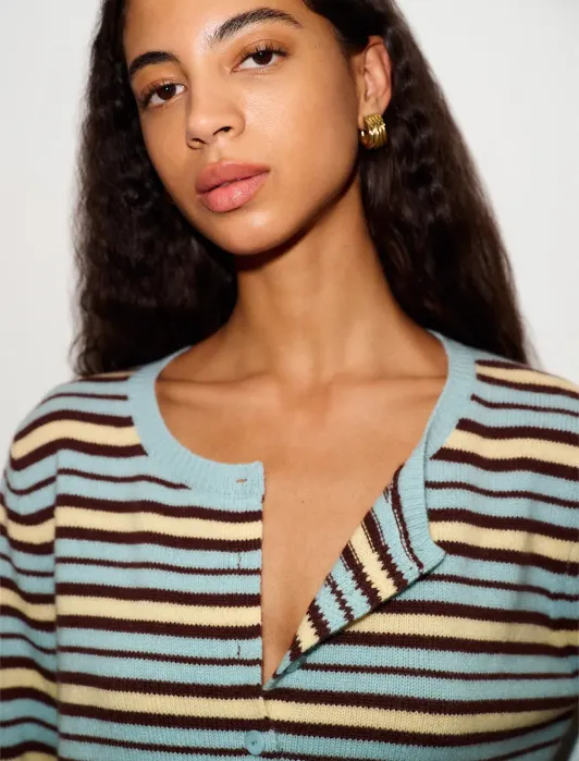Woman with long curly hair wears gold hoop earrings and a striped cardigan in blue, cream, and brown tones. The top button of her cardigan is undone. She has a neutral expression.