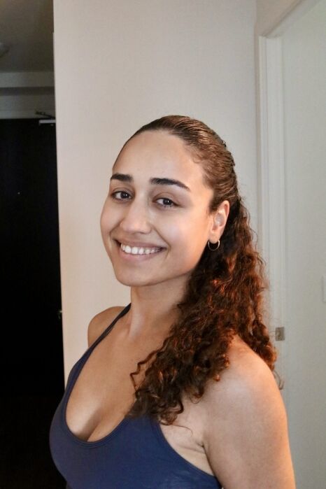 Woman with curly hair wearing a navy tank top smiles while standing indoors near a white wall and doorframe.