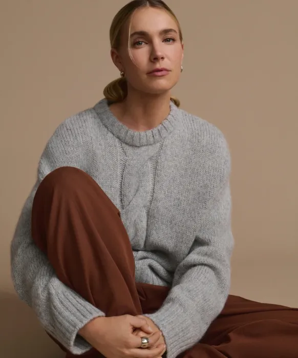 Woman with blonde hair in a low ponytail wears a gray knit sweater and brown pants, sitting against a beige background with a neutral expression.