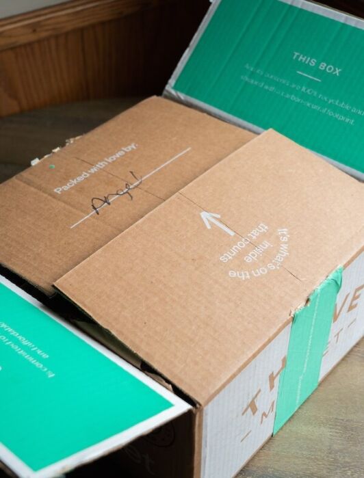 A partially open cardboard box with green flaps, showing an arrow and the text "Packed With Love By: Angel" on the top.