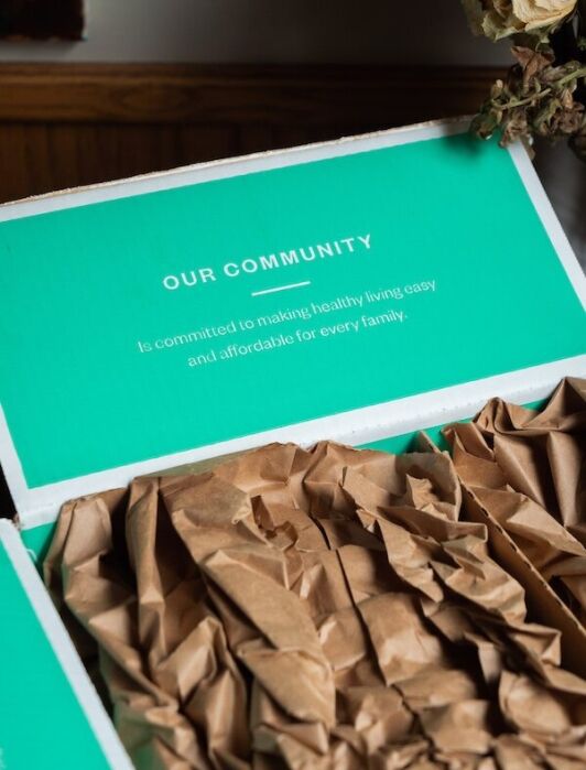 Open box with brown packing paper inside; the lid displays the text "Our Community is committed to making healthy living easy and affordable for every family.