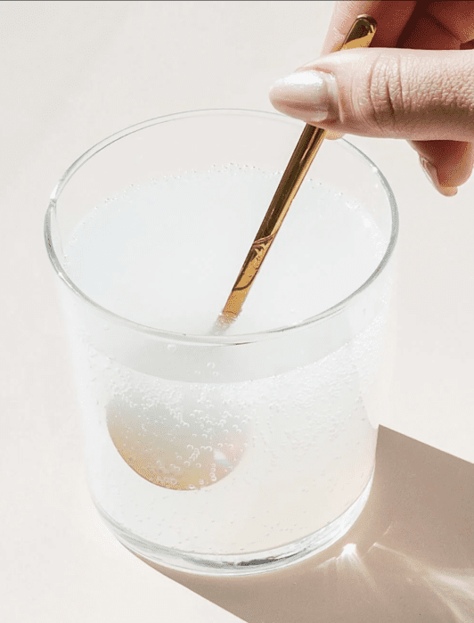 A hand stirs a bubbly white liquid in a clear glass with a golden spoon on a light surface.