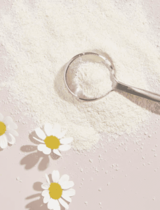 A metal scoop of white powder is on a pink surface with scattered powder and three white daisies.
