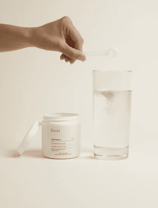 A hand holds a small scoop over an open jar next to a glass of clear liquid on a beige background.