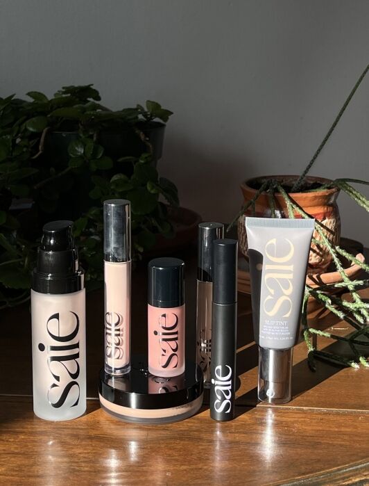 Collection of Saie cosmetic products arranged on a wooden surface, including bottles, tubes, and a compact, with plants in the background.