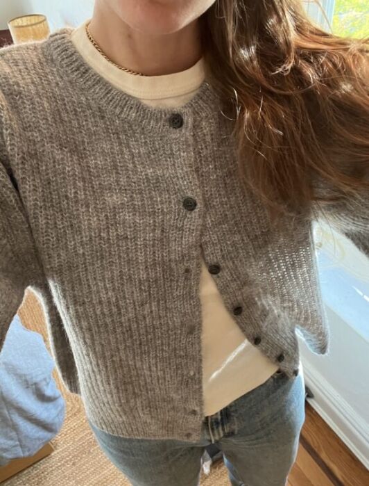 A person wearing a grey knit cardigan over a white shirt and blue jeans, standing indoors near a window.
