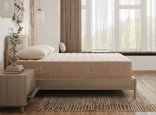A minimalist bedroom with a neatly made bed, light wood furniture, a nightstand with a vase, and large windows with sheer curtains letting in natural light.