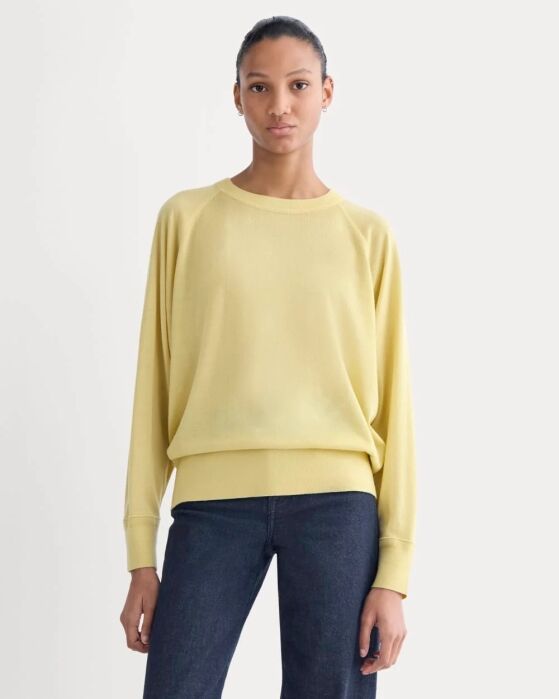 A woman stands facing forward, wearing a loose-fitting yellow sweater and dark blue jeans against a plain light background.