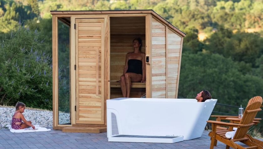 A woman sits in a small outdoor wooden sauna, while a man relaxes in a modern white bathtub and a child plays nearby, all set on a patio with trees in the background.