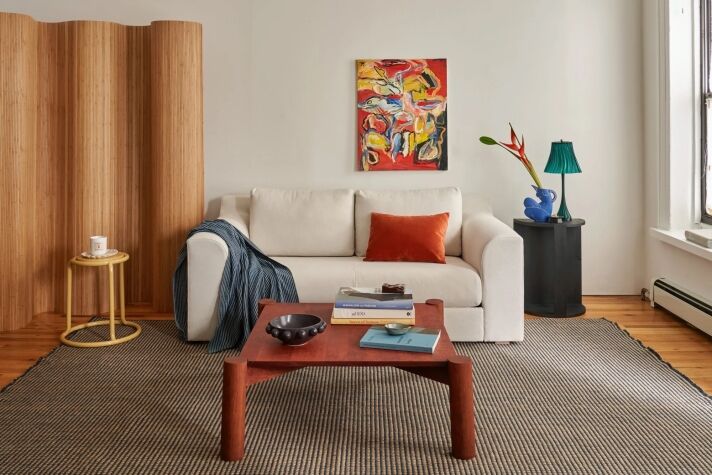 A modern living room with a white sofa, orange pillow, abstract wall art, wooden coffee table, side tables, a lamp, and decorative objects on a patterned rug.