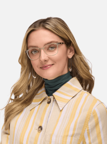 Woman with long blonde hair wearing clear eyeglasses, hoop earrings, a green turtleneck, and a yellow striped jacket, facing forward and smiling slightly.