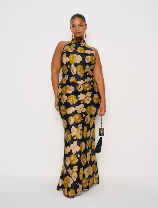 A woman stands against a white background wearing a sleeveless, floor-length black dress with a yellow and beige floral pattern, holding a small black bag.