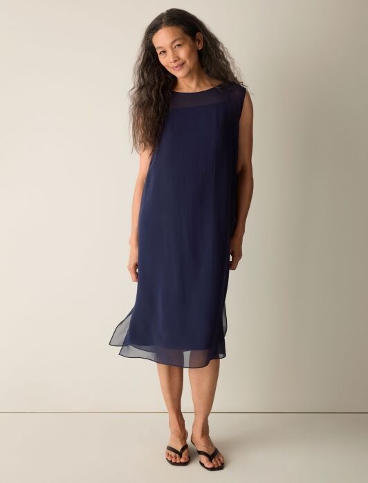 A woman stands against a plain background wearing a sleeveless, knee-length navy blue dress and black flip-flops.